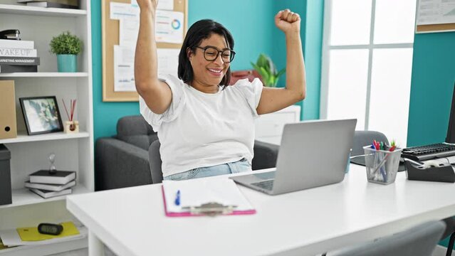 A middle-aged hispanic woman celebrates success in a modern office setting.