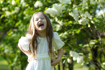 Cute happy little girl in a white dress is laughing in a spring park near the white lilac flowers....