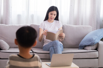 Cute Asian preschool boy has fun enjoys playing colorful toys alone in living room with mother, female mom reading book sitting on sofa relaxing with little kid after homeschool teaching using laptop