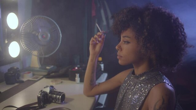 Afroamerican young woman tasting a cigarette in front of an old fashioned mirror, wearing a glitter dress.