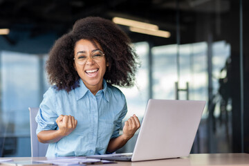Portrait of a happy and successful young business woman sitting in the office in front of the laptop and looking happy at the camera, celebrating and showing a victory gesture with her hands. - obrazy, fototapety, plakaty