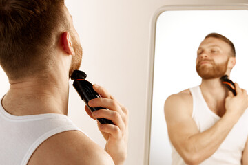 Serious man shave beard for hygiene, cleaning and hair removal with electric razor in front of...