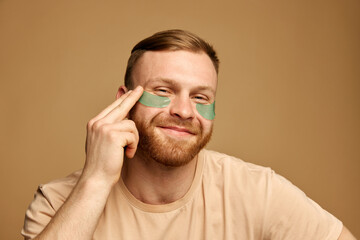 Fototapeta premium Portrait of overjoyed young man with beard applying hydrogel patches with moisturized effect under eyes against beige background. Concept of beauty treatment, selfcare and hygiene, cosmetic, spa.