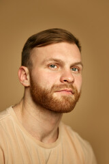 Fototapeta premium Portrait of young handsome redhead man with beard in beige T-shirt looking away against sandy studio background. Concept of beauty treatment, selfcare and hygiene, cosmetic, spa. Ad