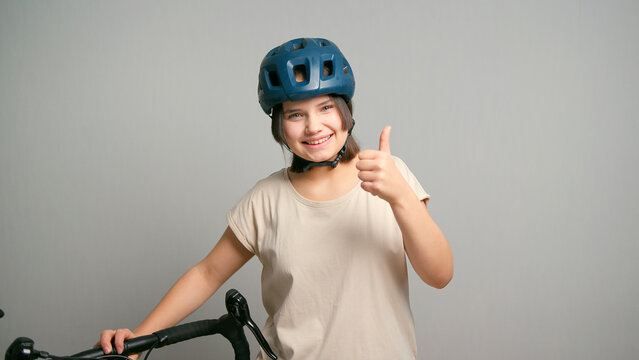 Young latin teenager female student with bicycle and blue safety bike helmet showing thumbs up smiling isolated on gray background. Copy space