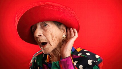 Funny portrait of smiling happy crazy elderly woman with no teeth, puts hand to ear to listen to a...