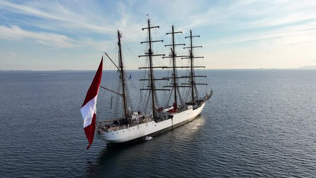 Aerial drone video of beautiful three mast barque or barc type classic sailing wooden boat with huge waving flag anchored in Mediterranean deep blue Aegean sea at sunset with golden colours