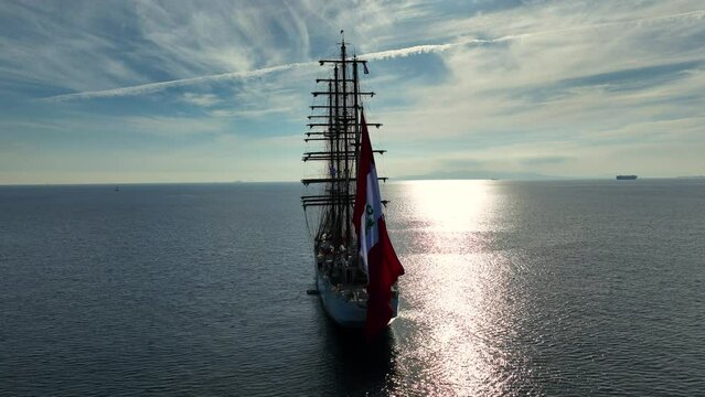 Aerial drone video of beautiful three mast barque or barc type classic sailing wooden boat with huge waving flag anchored in Mediterranean deep blue Aegean sea at sunset with golden colours