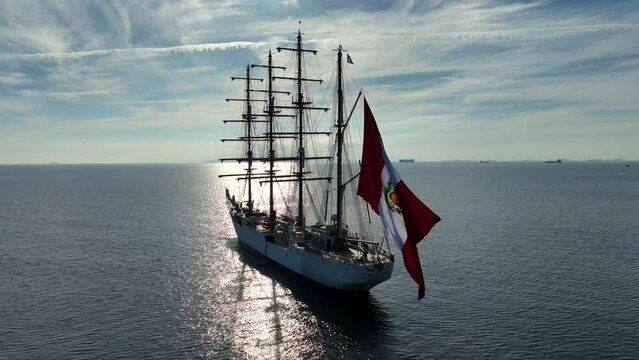 Aerial drone video of beautiful three mast barque or barc type classic sailing wooden boat with huge waving flag anchored in Mediterranean deep blue Aegean sea at sunset with golden colours