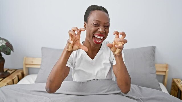 Smiling african american woman lying in bedroom, pulling a funny yet sexy claw gesture, exuding aggressive expression in comfort of home