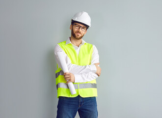 Man architect, construction engineer, builder, or foreman in white office shirt, yellow uniform workwear vest, safety hardhat, and glasses holding paper blueprint plan and standing by grey house wall