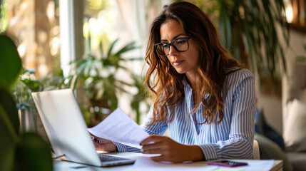 Close-up of a professional woman with long hair and glasses, wearing a blue striped shirt, reviewing papers with a laptop open in front of her, in a home office environment