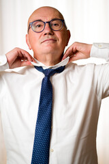 Elderly, bald, beardless man in trendy blue glasses, tying a bright tie with a subtle dot design on...