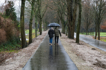 Bewegung bei schlechtem Wetter: Frau und Mann in Outdoorkleidung mit einem großen Regenschirm gehen im Dauerregen durch eine Allee auf einem asphaltiertem Weg spazieren, viel Copy Space
