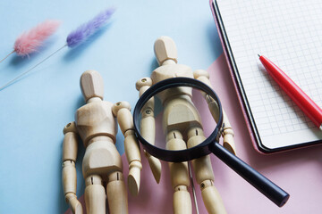 Wooden mannequins of a man, a magnifying glass and a notebook on a pink and blue background....