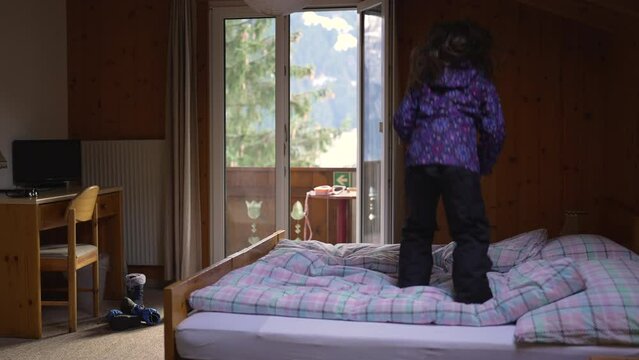 One happy little girl bouncing in bed inside Swiss wooden chalet during winter season in Switzerland. Carefree child jumping up and down