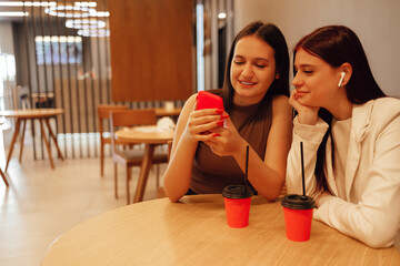 Two young girls are sitting in cafe with modern interior. Smiling longhaired brunettes women looking at smartphone at restaurant.