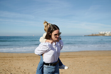 Young mother carries her little daughter on her back, walking along the beautiful waves breaking on the Atlantic shore