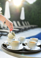 Woman's hand pouring cream into cups of coffee. In the background there is a swimming pool and sun loungers