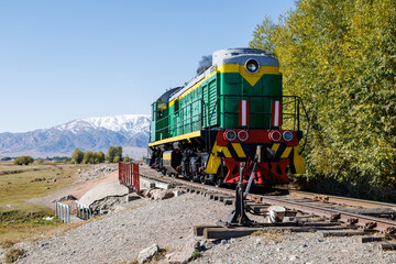 green diesel locomotive crossing small bridge with mountains in the background at summer day in...