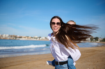 Caucasian happy young mother smiling looking at the camera while giving piggyback ride to her...