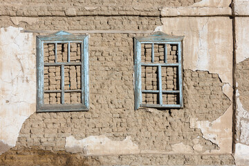 old house adobe air brick wall with two embedded wooden window frames and peeled off plaster leftovers - flat texture and full frame background.