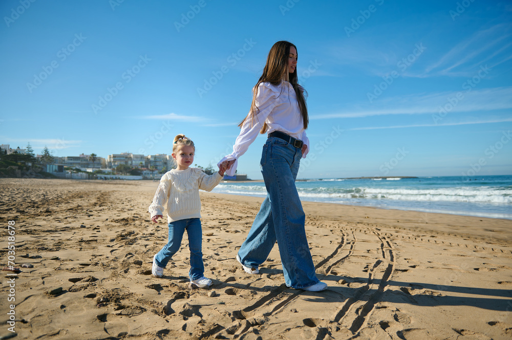 Wall mural young mother and daughter holding hands and walking along the tropical beach, enjoying a happy weeke