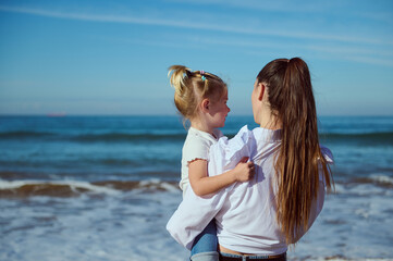 Rear view of mother holding her daughter and admiring waves splashing while pounding on the sandy shore. Family pastime