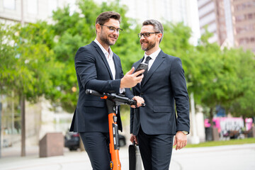 Two handsome young businessman using phone and standing on the street outdoor. Portrait of a smiling businessmen in formal suit standing outside office building using mobile phone.