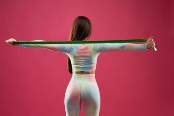 Young dark haired woman doing rubber band exercises indoors in studio. Back view of fitness slim female with spread arms, exercising with green elastic band, isolated on pink. Sport, health concept. - obrazy, fototapety, plakaty