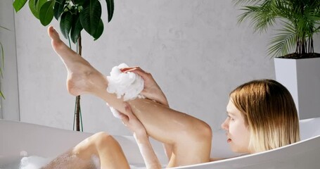 Focused girl washing neat legs, chilling in comfortable bathtub in luxury hotel. Side view of beautiful caucasian female scrubbing body with shower sponge, in front of grey wall. Self care concept.