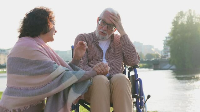 Sad old handicap man in wheelchair walking with caregiver senior woman in park. Elderly family couple woman supporting taking care of paralyzed man in chair for disabled people outdoor. Rehabilitation