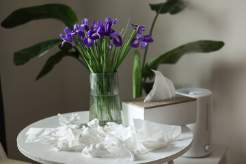 Seasonal allergy rhinitis to flowers. Bouquet of purple iris flowers in vase on table, many used napkins and humidifier or air purifier at home interior. Allergic symptom of odor pollen flowering