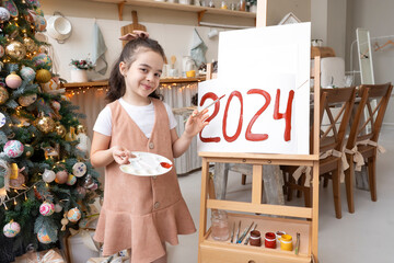 Pretty schoolgirl artist paints red numbers 2024 on easel with palette and brush. Christmas tree background. Art education at school. Merry Christmas and Happy New Year Eve concept.