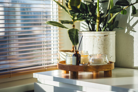 Apartment decoration with aroma reed diffuser, burning aromatic candles on wooden tray. Hygge, home aromatherapy, relaxation concept. Green plants on background. Bali oriental style in modern interior