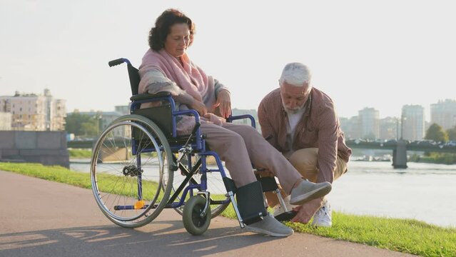 Senior handicap woman in wheelchair walking with caregiver old man in park. Elderly family couple man supporting taking care of paralyzed woman in chair for disabled people outdoor. Rehabilitation