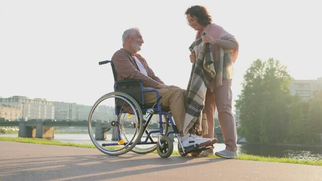 Old handicap man in wheelchair walking with caregiver senior woman in park. Elderly family couple woman supporting taking care of paralyzed man in chair for disabled people outdoor. Rehabilitation