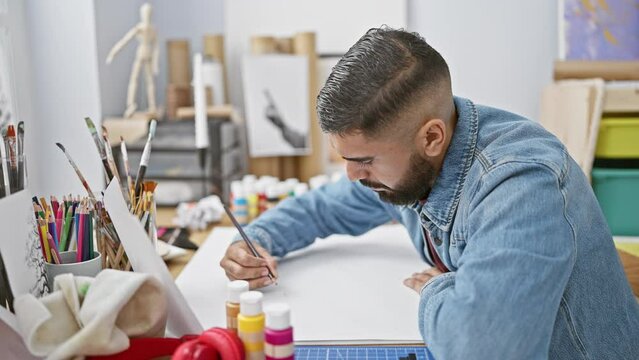 A focused man with a beard drawing in an art studio, surrounded by paint and pencils.