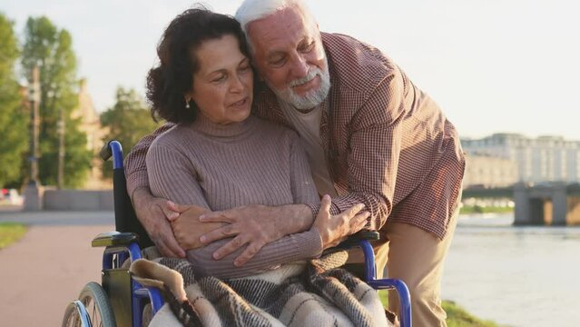 Senior handicap woman in wheelchair walking with caregiver old man on road in park. Elderly family couple man supporting embracing paralyzed woman in chair for disabled people outdoor. Rehabilitation