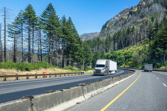 Two big rig semi trucks with refrigerator semi trailers running in opposite directions on the divided highway road with rock mountains and green trees on the sides