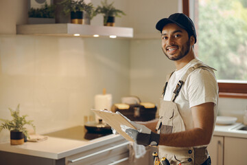 Smiling handsome handyman in uniform holding clipboard and looking at camera in kitchen