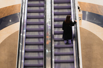 Faceless woman back view in a black coat standing on an escalator going up the stairs in a shopping...