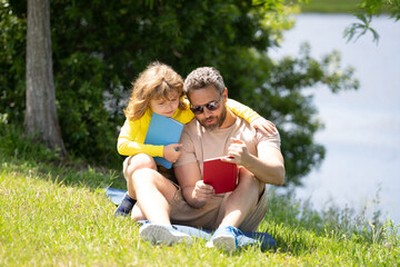 Fototapeta premium Father reading book with son in park, preparing school homework together, parenting. Summer lifestyle. Parenting and childhood concept. Little boy learning with father in outdoors garden.