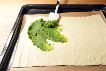 Raw puff pastry dough on baking paper and a tray is spread with homemade green pesto to bake savory...