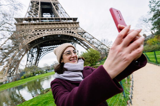 Happy young tourist woman using mobile phone to take selfie portrait with Eiffel Tower of Paris on the background. Travel and vacation concept.