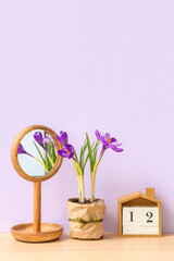 Pot with beautiful crocus flowers, mirror and cube calendar on table near lilac wall