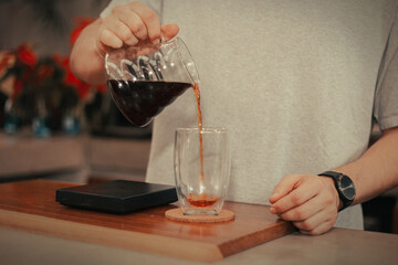 a person pouring coffee from a carafe into a clear mug on a wooden table
