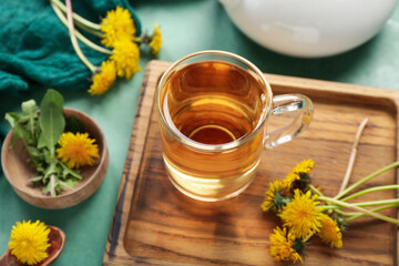 Board with glass cup of healthy dandelion tea on green background