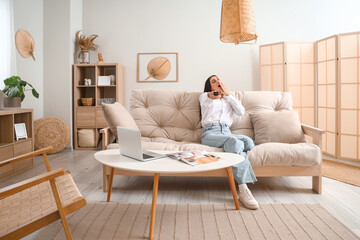 Sleepy young woman watching TV on couch in living room - obrazy, fototapety, plakaty
