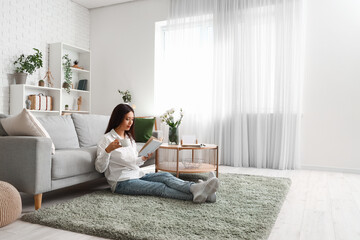 Beautiful young Asian woman with cup of tea reading book in living room - Powered by Adobe